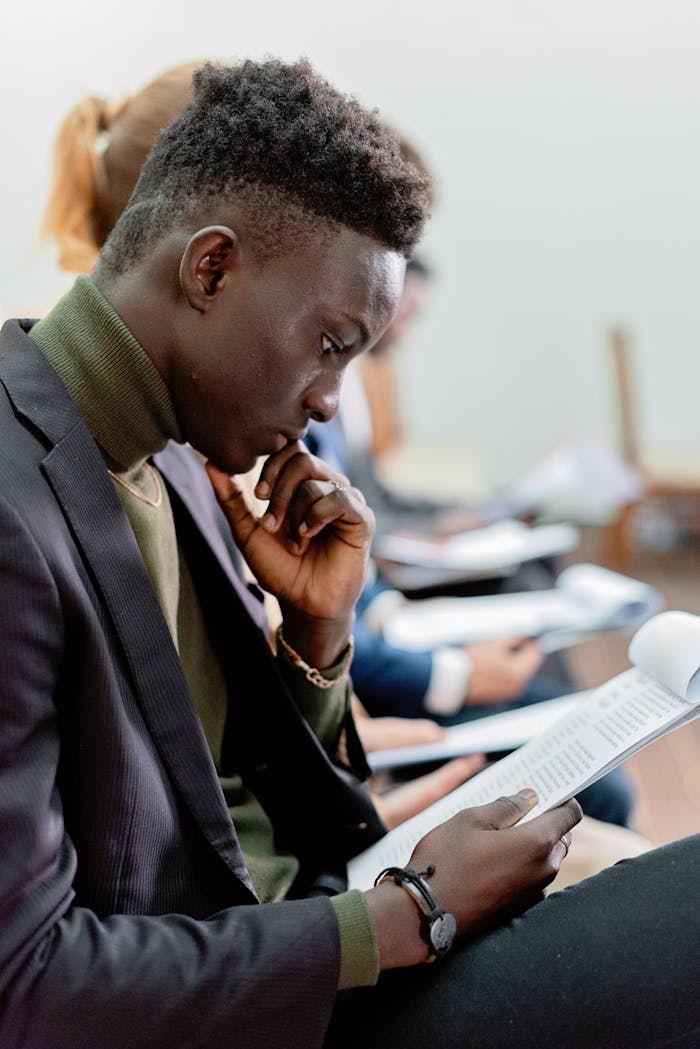 A young man in formal attire intently reads a document, surrounded by others in a similar setting, highlighting concentration and professional atmosphere.
